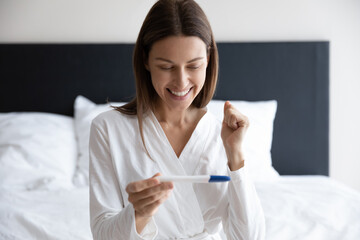 Overjoyed young woman sit on bed hold ovulation test feel excited getting pregnant, happy healthy female celebrate positive pregnancy confirmation results at home, maternity, fertility concept