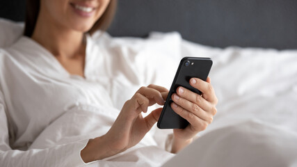 Close up of smiling young woman lie relax in white bed wake up in the morning using modern smartphone gadget, happy millennial female rest in hotel or home bedroom browsing Internet on cellphone