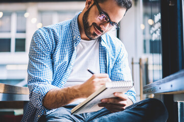Talented male designer drawing sketch in notebook while spending time in cafe interior, creative writer in spectacles making notes of journalistic article in notepad satisfied with occupation - obrazy, fototapety, plakaty