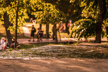 scene of a puddle of water on front and people walking in the park behind blur background. 