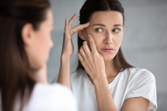 Anxious young woman look in the mirror worried about wrinkle or acne on unhealthy skin, upset unhappy millennial female examine squeeze pimple on face, cosmetology, skincare concept