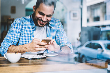 Fototapeta premium Happy hipster guy smiling reading message about winning online contest on smartphone resting in cafe,cheerful male person excited with receiving good news on telephone reading text enjoying coffee