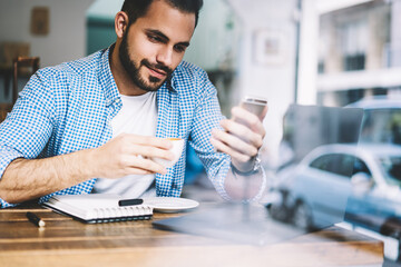 Bearded young hipster guy reading news from networks via smartphone sitting at coffee shop,male freelancer working in cafe downloading multimedia on telephone using application and 4G connection