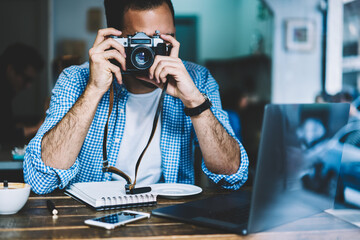 Skilled male photographer making picture of cafe sitting at table with netbook for editing,hipster...