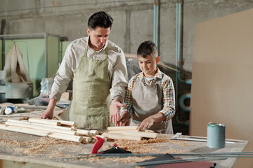 Male carpenter in apron pointing at planks while explaining teenage son how to work with wood in furniture workshop