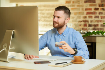 A man with a beard sits in front of the computer and holding a credit card at his hand at home. A guy doing an online payment on the internet on a desktop computer in his apartment. 