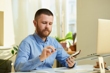 A man with a beard flipping the credit card near a computer at home. A guy in a blue shirt doing an online payment on a laptop in his apartment.