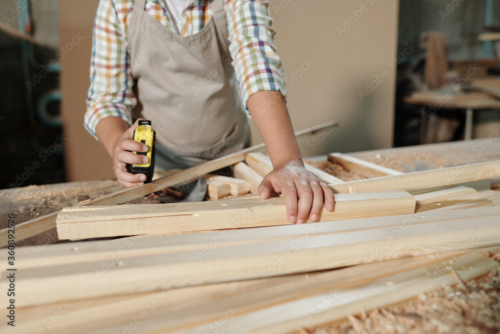 Wall mural Close-up of unrecognizable boy in apron using tape measure while working with wooden details in joinery workshop