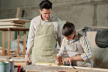 Middle-aged carpenter in apron standing at desk and watching how his teenage son making mark on plank in workshop