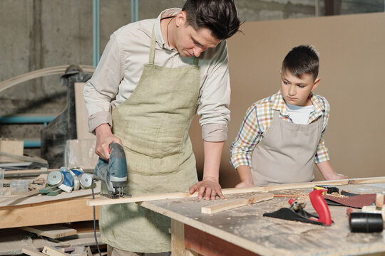 Busy skilled father in apron cutting wooden plank with jigsaw while his teenage son watching it in workshop
