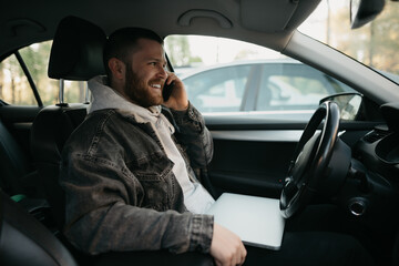 A happy man with a beard doing business calls on his smartphone inside a car, a laptop lies on his lap. A guy smiling stopped his car to immediately remotely solve tasks at work in social distance.