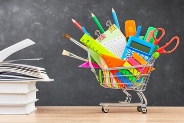 Various office supplies for school in a cart and textbooks on a wooden table on a blackboard...