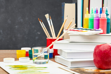 A textbook, acrylic and watercolor paints, brushes in a stand and a red Apple on a wooden table...