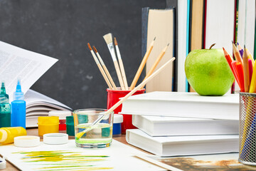 A textbook, acrylic and watercolor paints, brushes in a stand and a green Apple on a wooden table...
