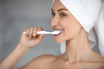 Smiling millennial woman in bath towel on head look in mirror brush healthy teeth with whitening toothpaste, happy millennial female perform daily facial routine in bathroom, oral hygiene concept