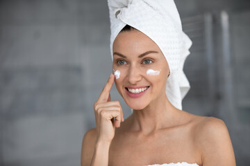 Young lady after shower look in mirror apply facial peeling or cream for healthy glowing skin,...