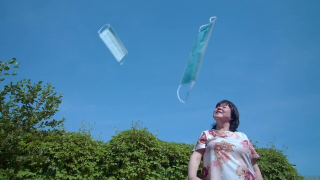 Girl happily throws up medical protective masks up in blue sky. One of masks flies right into camera. Shooting on summer sunny day against background of green foliage. Quarantine and disease exemption