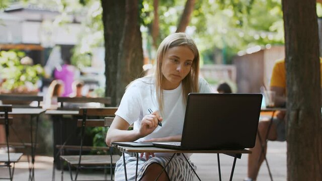 Young woman designer sitting at table on a summer terrace of cafe and drawing on digital tablet, using laptop. Sunny day. Concept of freelance