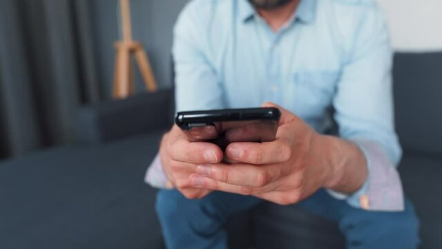 Formally dressed man sits on a sofa and using smartphone for texting message. Close-up