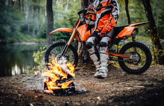 Charming young female racer wearing motocross outfit on her bike and warming up next to a bonfire in the woods. Cropped image