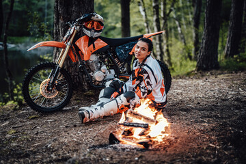 Beautiful young female racer wearing motocross outfit, warming up sitting next to the bonfire