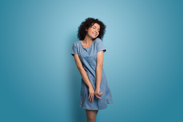 Delightful caucasian woman with curly hair wearing a blue summer dress is smiling on a studio wall