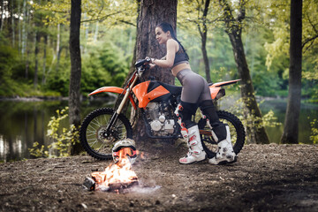 Beautiful young female racer wearing motocross outfit with semi naked torso leans her motorcycle on a tree next to a bonfire in the forest