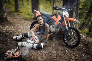 Beautiful young female racer wearing motocross outfit with semi naked torso, warming up while sitting next to the bonfire