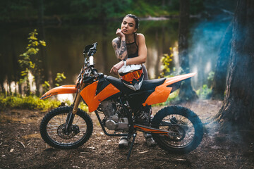 Plakat Sensual tattooed racer girl wearing motocross outfit with semi naked torso leaning on her bike and looking on camera in the woods