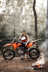 Fototapeta na wymiar Sensual tattooed racer girl wearing motocross outfit with semi naked torso leaning on her bike and looking on camera in the woods