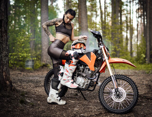 Fototapeta na wymiar Sensual tattooed racer girl wearing motocross outfit with semi naked torso posing next to her bikein the woods