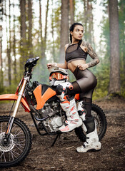 Naklejka na ściany i meble Sensual tattooed racer girl wearing motocross outfit with semi naked torso posing next to her bikein the woods