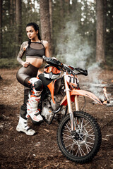 Sensual tattooed racer girl wearing motocross outfit with semi naked torso posing next to her bikein the woods