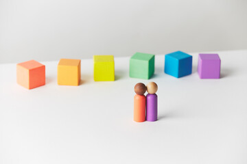 Wooden figurines concept. Couple standing in front of wooden blocks in colours of the gay pride rainbow flag. Couple of mixed skin colour. 