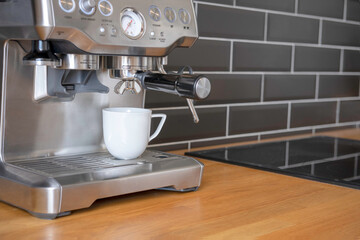 A white cup stands in a coffee machine in the process of making coffee in the kitchen with a black tile wall and a wooden tabletop at home. Morning ritual.