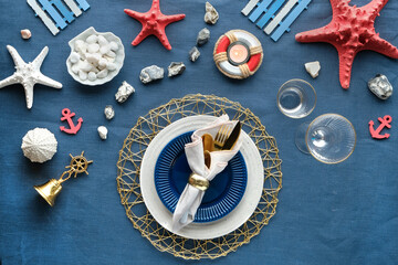 Columbus Day or Summer birthday lunch table, blue white with red. Flat lay, contemporary nautical table setting with sea decorations on classic blue linen textile. Plates, golden cutlery, utensils.