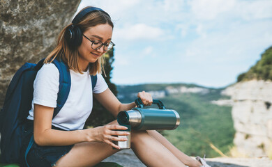girl traveler enjoys music with headphones and drinks tea from thermos while sitting in mountains...