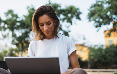 Stylish female entrepreneur working with modern laptop outdoor in summer at city park. Young professional woman looking monitor using tablet outside.
