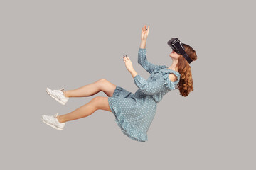 Gamer girl in ruffle dress hovering in air, levitating with virtual reality glasses on head, playing game through vr headset, floating in cyberspace. indoor studio shot isolated on gray background