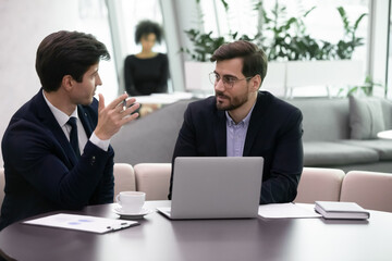 Young business man explaining to senior colleague new strategy. Serious advisor consulting client using computer, office manager listen to coworkers plan. Insurer or salesman giving advice to client