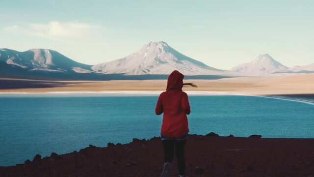 Hiker woman wearing a red hood running down the hill towards the lake Lejia. Aerial view of mountain lake panorama in the Atacama desert. 4k