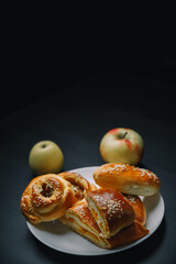 Vertical shot of delicious sesame buns or pies on white plate and yellow apple on black wooden table. Tasty unhealthy snack for lunch. Overweight, diet, nutrition, eating habits concept. Copy space.