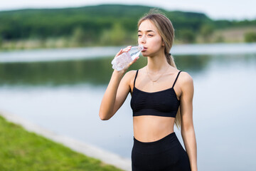 Fit beautiful young woman drinking water after workout by the lake. Teenage runner at the beach with bottle of water refreshing after workout. Fitness, diet,active lifestyle concept.