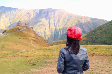 Naklejka premium Female Traveler Enjoying the Hilltop Gergeti Trinity Church with Kazbek Mountain in the Backdrop, Stepantsminda, Georgia