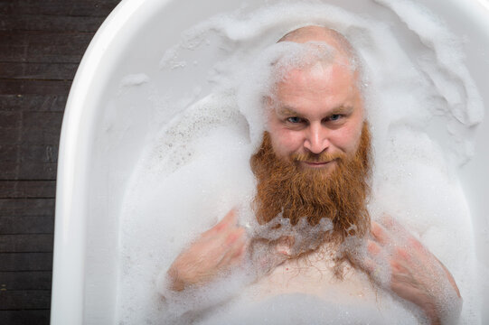 A Bald Man Is Fooling Around In The Foam Bath. The Guy Makes Funny Hairstyles On His Red Beard.