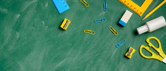 Blue and yellow school stationery on green chalkboard. Flat lay, top view, overhead. Modern student workspace. Back to school banner template, education concept.