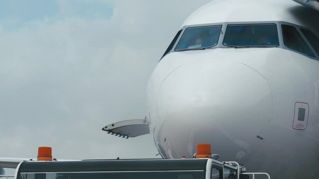 Close front outside view of airplane cockpit. Close-up of a white modern airplane at the airport in a parking lot against a cloudy sky. Service car with orange flashing lights at the airport.
