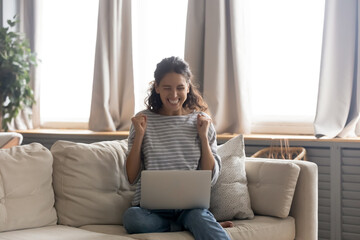 Excited young woman looking at laptop screen, celebrating win, showing yes gesture, sitting on cozy...
