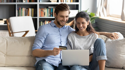 Happy young couple shopping together, customers paying by credit card online, sitting on cozy couch...