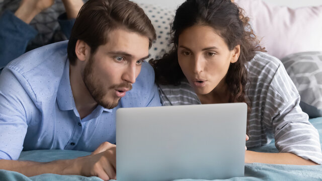 Interested Young Man And Woman Looking At Laptop Screen Together, Lying In Bed At Home, Puzzled Surprised Couple Reading Good News In Email, Great Shopping Offer, Watching Video In Social Network